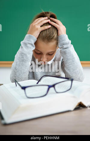 Sad girl holding her head. Photo of schoolgirl with book on the desk in classroom. Back to school Stock Photo