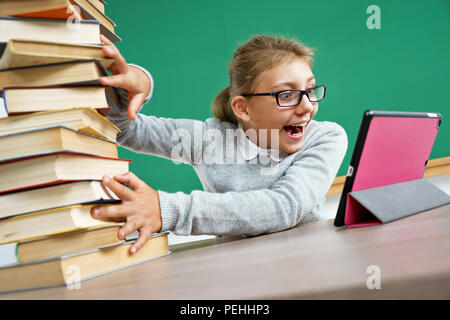 Happy girl looking at the tablet in the same time pushes off the mountain of books. Photo of little girl in classroom. Education concept Stock Photo