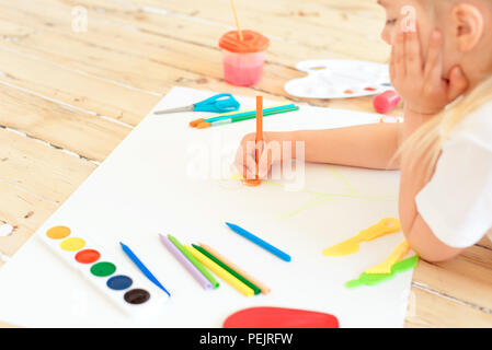 Little blonde girl painting on big white paper while laying on the floor indoors. Stock Photo