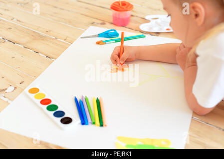 Little blonde girl painting on big white paper while laying on the floor indoors. Stock Photo