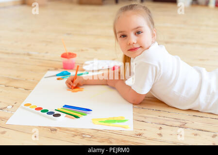 Little blonde girl painting on big white paper while laying on the floor indoors. Stock Photo