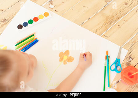 Little blonde girl painting on big white paper while laying on the floor indoors. Stock Photo