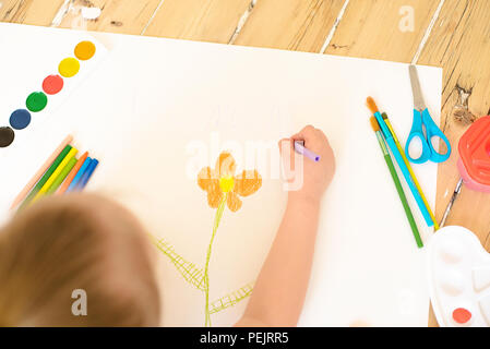 Little blonde girl painting on big white paper while laying on the floor indoors. Stock Photo