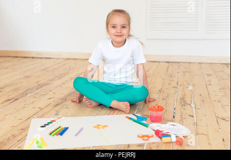 Little blonde girl paintson big white paper sitting on the floor indoors. Stock Photo