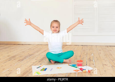 Little blonde girl paintson big white paper sitting on the floor indoors. Stock Photo