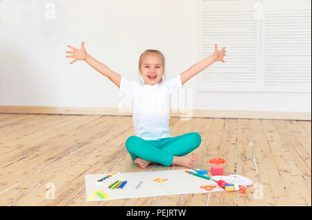 Little blonde girl paintson big white paper sitting on the floor indoors. Stock Photo