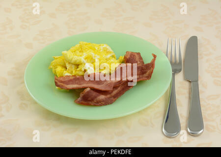 Quick breakfast of scrambled eggs and two strips of turkey bacon on green plate with fork and knife Stock Photo