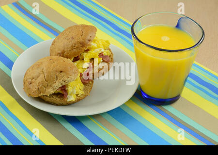 Breakfast sandwiches of scrambled eggs with pieces of turkey bacon on whole wheat rolls and glass of orange juice Stock Photo