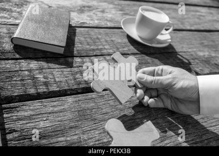 Greyscale close up on hand of unidentifiable business man holding puzzle piece over rustic wooden table for concept about solutions to problems. Stock Photo