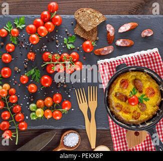fried omelette from chicken eggs with red cherry tomatoes and smoked sausage in a round black frying pan , top view Stock Photo