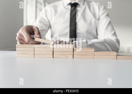 Close-up view of incognito man in white shirt building little wall or staircase of wooden bricks over office table. Stock Photo