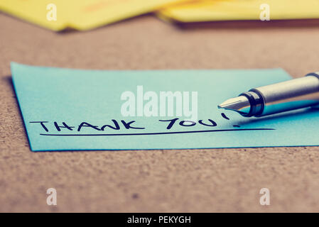 Handwritten Thank You note written on a blue sticky note lying on a cork board with a fountain pen viewed low angle. Stock Photo