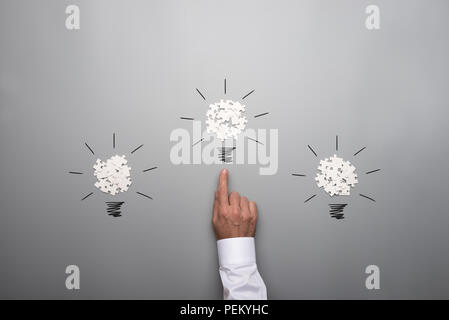 Three light bulbs formed by scattered white puzzle pieces with hand of a businessman pointing towards the bulb in the middle. Over grey background. Stock Photo