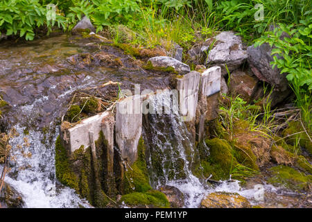 A small waterfall between the stones and two wooden planks among the green grass from a creek with clear mountain water in the mountains of the Altai Stock Photo