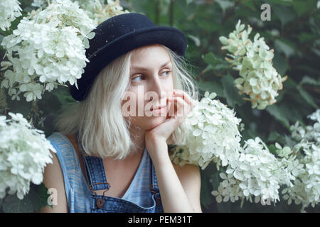 Closeup portrait of beautiful Caucasian teenage young blonde alternative model girl woman in blue tshirt, jeans romper, black hat, sitting among large Stock Photo