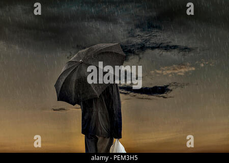 Man In The Rain.  Man holding a black umbrella in the rain. Dramatic sky at dawn . Stock Photo