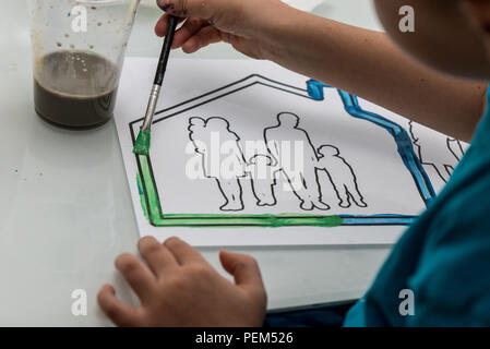 Young child colouring in a sketch of a family in a house using a paintbrush and watercolour paints in an over the shoulder view of the artwork. Stock Photo