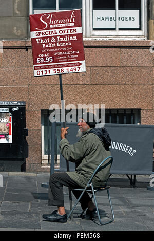 An mature Asian man sitting on the Royal Mile and holding a sign advertising a Thai and Indian restaurant in Edinburgh, Scotland, UK. Stock Photo