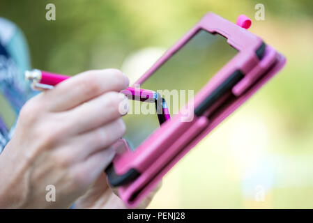 Close up view of the hand of a woman standing outdoors writing on the touch screen of a pink tablet with a stylus. Stock Photo