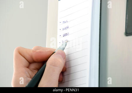 Female hand writing grocery list on a sticky reminder note on a refrigerator. Stock Photo