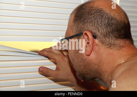 The bald man presses the rails of the blinds and looks out the window. Stock Photo