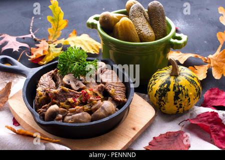 fried meat with mushrooms in a frying pan. on a black background. Lunch with autumn leaves. Top view Stock Photo