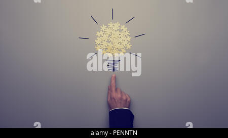 Retro toned image of a businessman pointing on a light bulb drawing with yellow puzzle pieces over grey background. Stock Photo