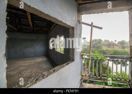 An empty room can be seen through the window of a cement building, palm trees and water can be seen in the background. Ganta Liberia Stock Photo