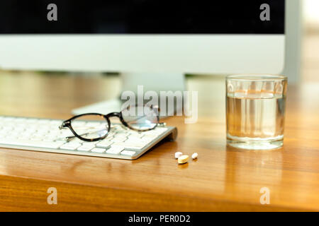 Conceptual work stress glass of water and pills on wooden desk with PC and glasses.
