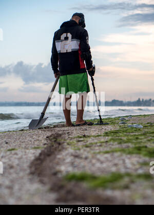 Path of a shovel behind a man searching for a precious metal using a metal detector on lonely beach at sunset time Stock Photo