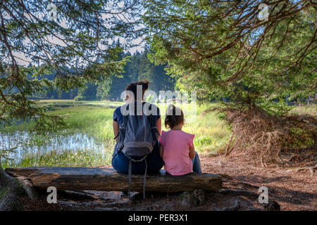 Mother and daughter sitting on bench near mountain lake, facing away, family outdoor hiking in nature, love between mothers and daughters Stock Photo