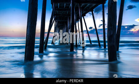 Amazing long exposure of sunrise under a wooden ocean pier. Smooth fuzzy blur created by the camera with a dramatic sky on the horizon. Stock Photo