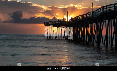 The first fishermen of the day on the end of an ocean pier as the sun rises. Sun beams explode from behind clouds and a bright orange glow. Stock Photo