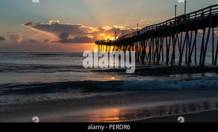 A long wooden fishing pier extends far into the ocean. The sun rising on the horizon at the end of the pier. Sunbeams and light rays. Stock Photo