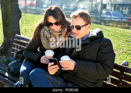 Young man and woman at cafe looking and using mobile phone. Young couple sitting in the park, drinking coffee to go, looking at his smart phone. Stock Photo