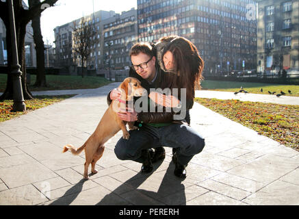 Girlfriend hugging young man while he crouching and cuddle the dog and all enjoy in a beautiful day in the park. Young happy couple with dog enjoy. Stock Photo
