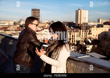 Couple enjoying the sun on the roof, young man is relaxed and girl holding coffee and looking at smartphone. Stock Photo