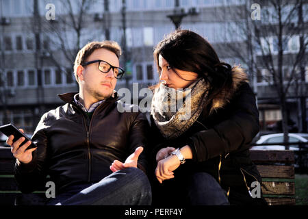 Couple on a park bench looking at each other, guy holding smartphone and the girl shows on her watch. Stock Photo