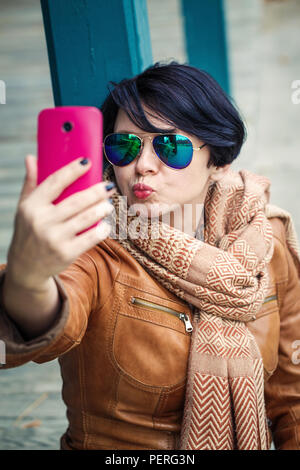 Portrait of middle aged caucasian brunette woman with short bob hair  in sunglasses wearing light brown leather jacket and scarf outside making selfie Stock Photo