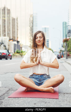 Portrait of slim fit sporty young white Caucasian hippie hipster woman with long blond hair meditating doing yoga exercisers in city street, healthy l Stock Photo