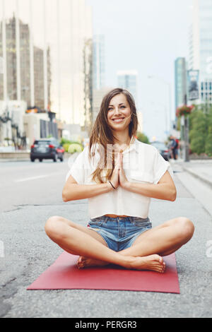 Portrait of slim fit sporty young white Caucasian hippie hipster woman with long blond hair meditating doing yoga exercisers in city street, healthy l Stock Photo