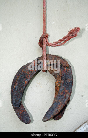 Rusted lucky charm horse shoe dangling on a red rope against a white wall bringing luck and averting disaster according to old wive tales. Stock Photo