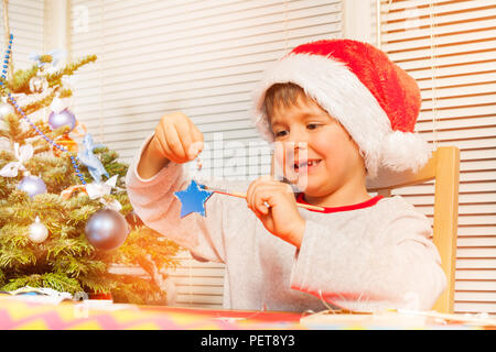 Portrait of preschool boy in Santa's costume holding wooden Christmas star and decorating it with blue paint Stock Photo