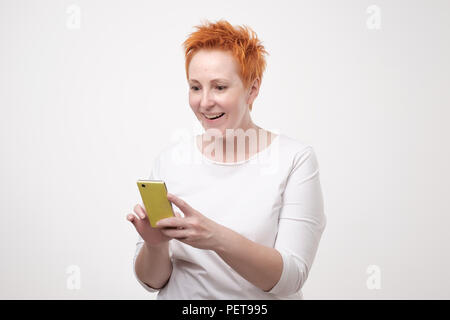 Close up portrait of businesswoman in white t-shirt and red short hair s looking at cell phone. She received interesting offer through sms Stock Photo