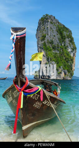 Portrait view of a traditional long-tail boat with Thai flags waiting on one of the beaches of Krabi in Thailand, with limestone cliffs in the back Stock Photo