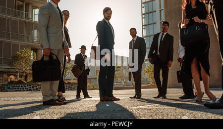 People standing on a busy city street with sun flare in the background. Men and woman in formal clothes commuting to office in the morning carrying of Stock Photo