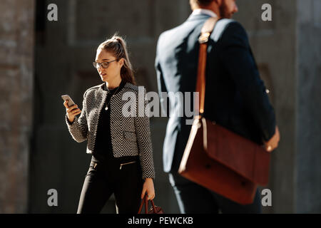 Businesswoman commuting to office in the morning looking at her mobile phone. Business people walking on city street to office carrying their office b Stock Photo