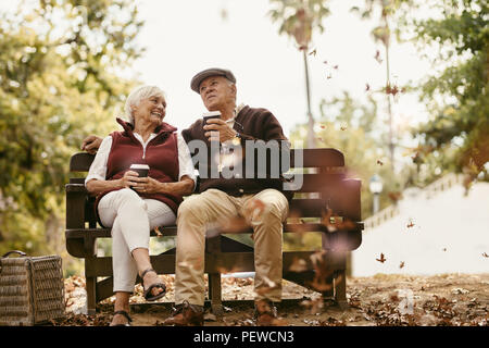 Relaxed senior couple on picnic sitting on park bench talking and having coffee. Old man and woman relaxing at park having coffee. Stock Photo