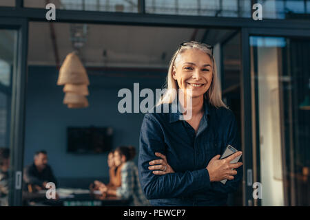 Portrait of smiling senior woman standing in office doorway with her arms crossed. Beautiful female executive at office with people meeting in backgro Stock Photo