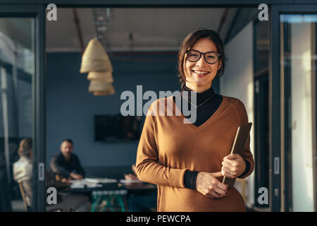 Portrait of positive female executive holding a laptop standing in office with colleagues working in background. Smiling business woman in casuals at  Stock Photo